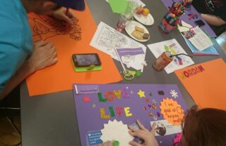 3 gda members lean over a table with bright orange and purple paper laid out, as they add colourful designs and texts to great art pieces.