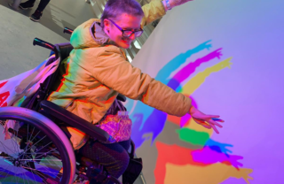 A GDA member in her wheelchair at the Science Centre strikes a dramatic pose, grinning with her arms held high in front of a prism of light - it casts many rainbow silhouettes against a plain white wall.