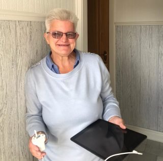 A woman with short hair and dark glasses stands in the hallway of her home, leaning against the wall, smiling and holding her new tablet in one hand and a the plug form its charger in the other hand.