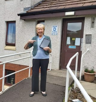 One of our members holds a tablet in her right hand, while standing outside her residence.