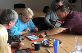 a group of people are sat around a table with late afternoon light coming in through the window. the group each have a tablet device and are discussing its use, with one member reaching over and pointing on the screen of another member's tablet.