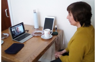a woman with shoulder length brown hair and a mustard yellow jumper sits profile at her desk, with a cup of tea, a laptop, biscuits and a bag on the table. She's facing and interacting with the group meeting taking place on her laptop.