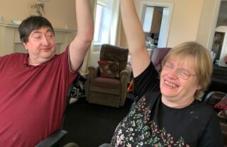 two people side by side in wheelchairs reach up their arms in a stretch, smiling towards each other. They are sat in a large lounge space.