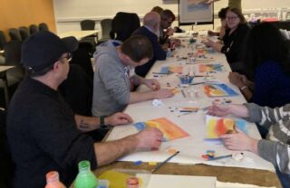 a long table is photographed from one end, stretching down the length of the room to a tutor who stands at a flipchart. A long roll of paper stretches the length of the table, and GDA members seated on either side work on a collective artwork.