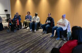 a grou pof 6 GDA members are seated in a hotel function room with their hands clasped and eyes closed in a peaceful relaxed pose. The room has wooden walls and the flooring is patterned in vertical stripes.