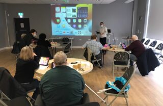 3 circular tables with GDA members sat at them fill a room with wooden floors and grey walls. Projected on the main wall is the screen from an iPad, while a tutor holding the iPad leads a presentation on how to operate it.