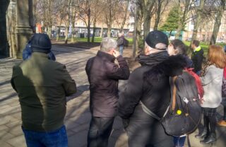 a group of 8 GDA members and a tutor are photographed from the back as they listen to the tour guide, outdoors on a sunny but cold looking day. The group are stood on a concrete pathway in a public park, with trees lining one side and the side of an old brick monument to the left.
