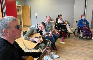 Five GDA members sit in a semi circle in a bright room with a wooden floor and grey walls. The two members to the left are playing acoustic guitars while everyone watches a tutor (out of shot) for direction.