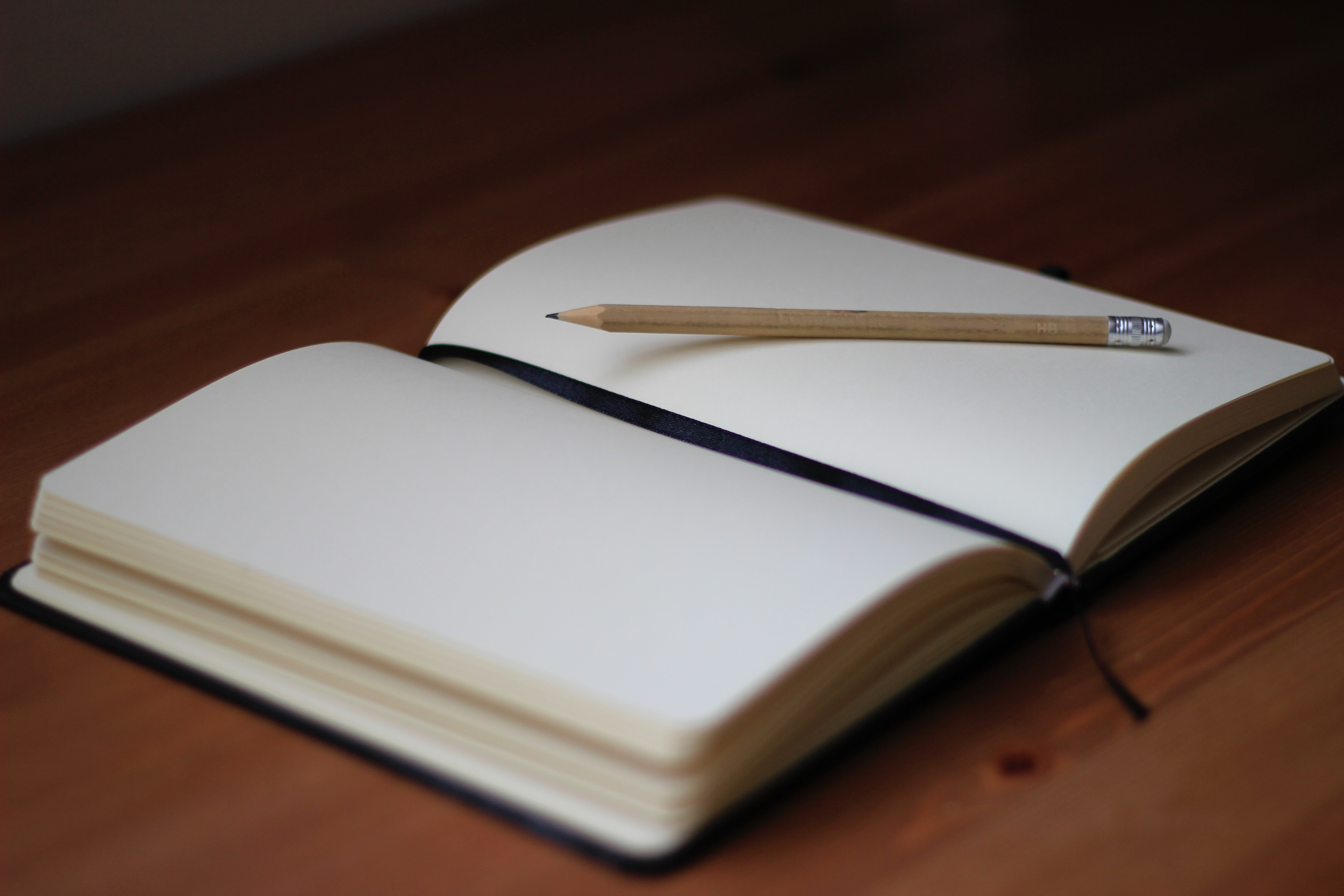 A notepad with blank white pages lays open on a wooden table. The book fills the frame, with a black ribbon marker running down the spine and a wooden pencil laying on the right hand page.