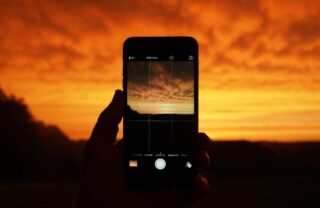 A phone screen is held up in someone's left hand on camera mode, to take a photograph of the sunset and clouds. The sky and clouds are a vivid orange and yellow.