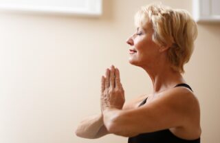 a woman with short blond hair sits profile in a room warmly lit with daylight. her body from the torso up is in shot and she is sat with her eyes closed and her hands together in a namaste pose