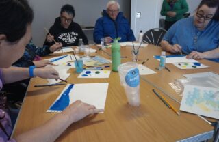 a group of GDA members sit in a semi circle around a table which takes up most of the shot. on the brown table are white sheets of paper and paints, with members concentrating, busy creating paintings.