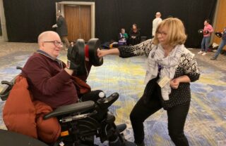 a gda member in a powerchair, red jumper and glasses, is on the left and wearing glove pads while a GDA staff member in a karate pose lands a punch on the pads with a comical grimace on her face