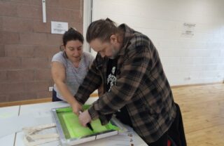 GDA member in a brown checked shirt in a well lit community hall leans over a table and presses the screen printing squeegee across the screen, pushing paint onto the paper. They are assisted by the tutor in a grey top.