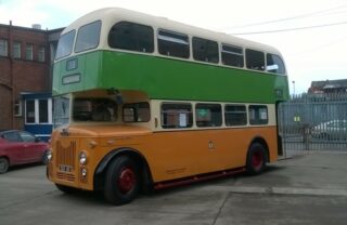 An old Glasgow double decker bus sits in the open courtyard of the garage, sat at an angle and facing the bottom left of the picture. The bus is a khaki green on the top level and a tangerine orange on the bottom, with old style lights, signage and deep red hubcaps on the wheels.