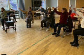 Members sit in a semi-circle of chairs and raise their arms into a ballet pose. The room is lit with afternoon light.