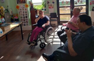 GDA members sit in a sunny room with windows behind them. They're seated on in wheelchairs and faced a computer screen. They smile and take part in the yoga exercises.