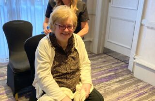 A member sits with a big, relaxed smile on her face, while a massage practitioner stands behind her massaging the top of her back.