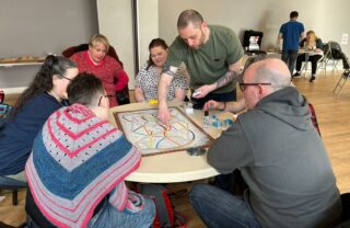 A group of members sit at a circular table with a colourful board game at the centre. A tutor leans over and points to the board, explaining the rules of the game.
