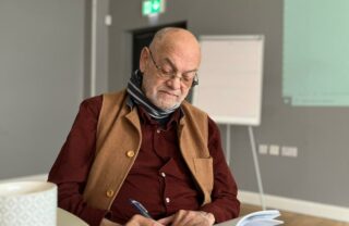 A GDA member in a deep red shirt and trousers with a brown bodywarmer on top sits with an A4 notepad open on his knee, looking down and writing with a pen.