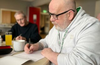 a GDA member sits at a table in the foreground with a white hoodie and glasses, writing on a notepad and looking thoughtful. Other GDA members sat at the table are visible in the background writing too.