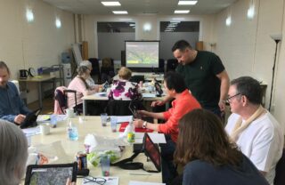 GDA members sit at 2 tables that lead towards a projector showing an iPad screen. Members have an iPad each and are learning new uses for them while a tutor moves around the room assisting people.