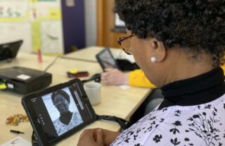 photo taken from over the shoulder of a GDA member as she sits looking down at her iPad sat on a table. The iPad displays a black and white portrait she has taken of herself smiling.