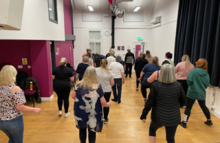 a large group of people in a community hall are line dancing, the image shows the group from behind in 4 rows, following the tutor's moves.