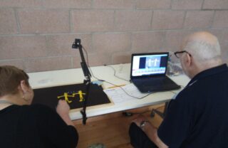 2 GDA members sit next to a laptop and a black slate, the slate has a small camera overhead and is capturing stop motion images of two yellow people figures and the word 'hello'.