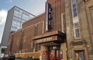 the outside facade of the GFT, featuring the GFT lettering vertical on a sign over the canopy entrance. The building is art deco in style, sat on a slightly sloped street on a bright day.