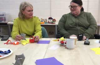 two GDA members sit at a table with sheets of paper and origami creations around them including small boxes, handbags and a fox. They are facing each other smiling and behind them are further origami creations on a table including a horse and patterned boxes.
