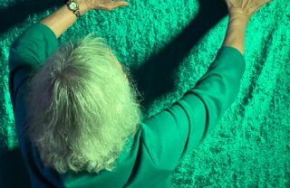 a GDA member stands with her back to the photographer. She is stood with her hands held up and to the right, casting dark shadows of her arms on a green felt backdrop. She's wearing a similar green colour of top which blends with the background while her white/grey hair contrasts against it.