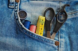 close up on the pocket of a pair of jeans. A pair of scissors, a yellow measuring tape, a button and 2 different colours of thread are poking out of the pocket.