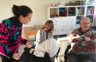 3 GDA members and a tutor are sat in a brightly lit room in a circle of chairs. One member smiles listening to the tutor while the others look off into the room.