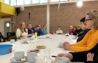 members sit around a long white table in a community centre with paper, cups of tea and biscuits and discuss comedy and funny stories. Members are smiling and interacting.
