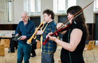 2 GDA members and a tutor stand in a brightly lit modern room in a row playing the keyboard, trumpet and violin together.