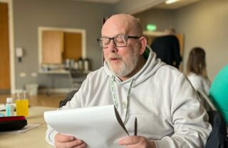 a GDA member sits in a white hoodie and blue jeans, facing the off to the side of the camera and holding a notepad. He's reading his writing from the notepad aloud with a slight smile.