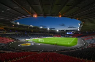 Hampden football stadium, a photo taken from inside the stadium in one corner of the seating. The sun sets through the gap in the stadium roof and the pitch is lit up in floodlights.
