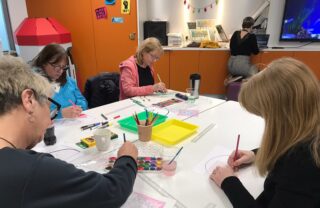 Four GDA Members are gathered around a square table in a classroom. They are each working on a drawing or a painting. Art materials are laid out on the table.