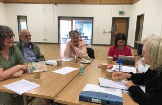 Six GDA members dressed in casual clothes are sat around a square table. They are indoors, in a hall, with windows behind them. On the table, there are paper coffee cups, A4 sheets of paper with notes, a ringbinder and an iPad. One of the members is talking to the whole group.