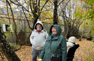 In the foreground, Two GDA members are standing in the woods. One is wearing a grey zip up hoodie and the other is wearing a green rain jacket. Both of them have their hoods up. They are standing in a clearing in the woods, and the ground is covered with yellow and orange fallen leaves. Behind them is another GDA member with their back to them in a power chair. They are wearing a beige rain jacket with their hood up. There is a canopy of bare trees surrounding them all.