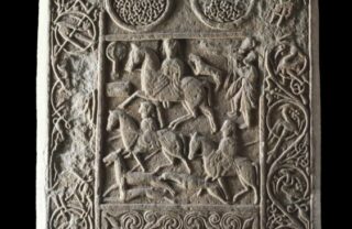 a large rectangular ancient stone is carved with celtic circular patterns and figures on horseback. The stone is grey in colour and is starkly lit from the side with a black background.