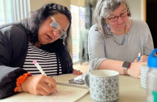 two GDA members are sat at a table in a community centre on a bright day with plenty light coming through the window off camera to the left. the photo is a close up of the two women busy writing on paper in front of them, with coffee cups, water and stationery in the table too.