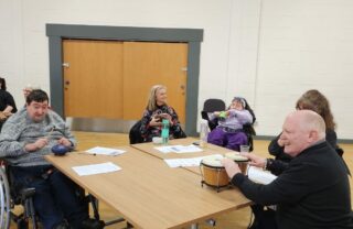 GDA members are sat round a square table smiling and playing on small percussion instruments.