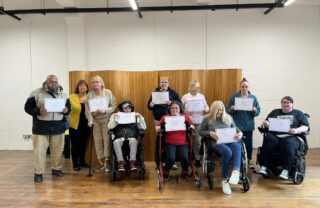 a group of GDA members stand or sit in a well lit room with a wooden backdrop. They are posing with their certificates from completing the confidence booster course and smiling.