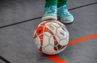 a close up of someone controlling a white and orange coloured football by putting their foot on top of it. Their trainers are light blue and the flooring is from a gym hall, with line markings.