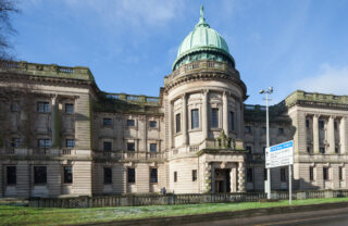 The Mitchell Library is sat against a blue sunny sky. The bricks are of light colour and the rounded roof turret has a green colour.