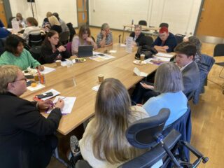 The photograph shows several GDA members sitting around a large table. In the middle of the near long side is Gordon McKee, MP for Glasgow South. He is turning around talking to the woman directly beside him.