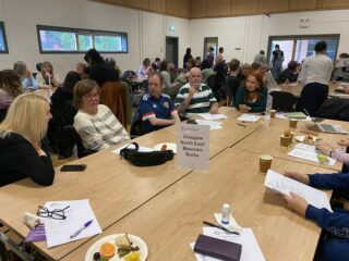 The photograph shows several GDA members sitting around a large table. At the far end of the table is a woman who is form the office of Maureen Burke, MP for Glasgow North East. She is listening to the other people at the table who are talking. In the background is the room crowded with other GDA members sitting at a number of tables.